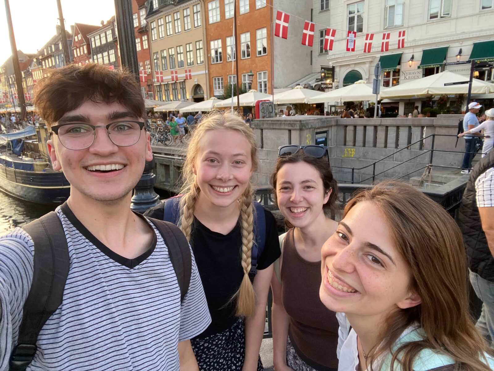 Four students posing near the edge of a canal, with a busy shopping street close behind them.